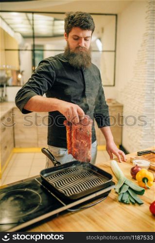 Bearded chef cooking meat in a pan on the kitchen. Man preparing boiled pork on table electric stove. Bearded chef cooking meat in a pan on the kitchen