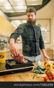 Bearded chef cooking meat in a pan on the kitchen. Man preparing boiled pork on table electric stove