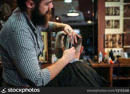 Bearded coiffeur cutting hairstyle by scissors. Client man in black salon cape sitting against a mirror at the barbershop