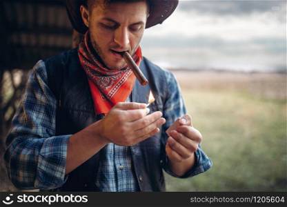 Bearded cowboy lights a cigar with matches, texas ranch on background, western. Vintage male person in leather clothes on farm, wild west culture. Bearded cowboy lights a cigar, wild west culture