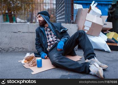 Bearded dirty beggar with food sitting at the trashcan on city street. Poverty is a social problem, homelessness and loneliness, alcoholism and drunk addiction. Dirty beggar with food sitting at the trashcan
