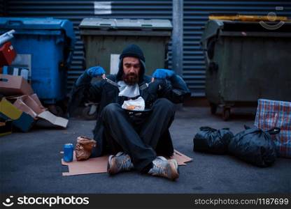 Bearded dirty beggar with knife and fork eats at the trashcan on city street. Poverty is a social problem, homelessness and loneliness, alcoholism and drunk addiction, urban lonely. Dirty beggar with knife and fork eats at trashcan