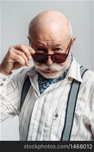Bearded elderly man with mustache poses in sunglasses, grey background. Mature senior looking at camera. Elderly man with mustache poses in sunglasses