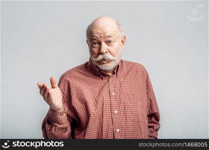 Bearded elderly man with mustache stands in a shirt, grey background. Mature senior looking at camera in studio. Bearded elderly man with mustache stands in shirt