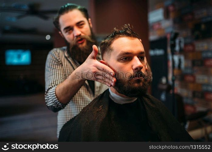 Bearded hairdresser making male hipster hairstyle. Client man in black salon cape visit barbershop