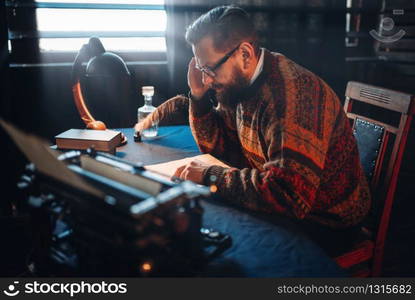 Bearded journalist in glasses reading his work. Retro typewriter, feather, crystal decanter, books and vintage lamp on the desk. Bearded journalist in glasses reading his work