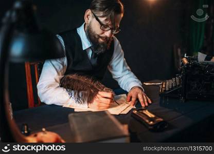 Bearded journalistr in glasses writes with a feather. Retro typewriter, books and vintage lamp on the desk. Bearded journalistr in glasses writes with feather