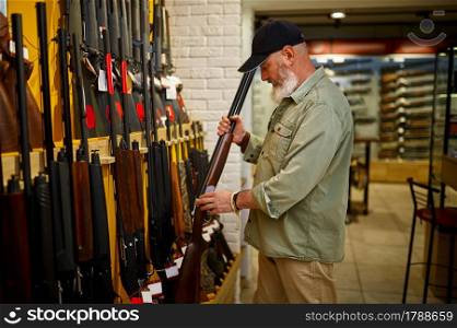 Bearded male hunter at the showcase with rows of hunting rifles in gun store. Weapon shop interior, ammo and ammunition assortment, firearms choice, shooting hobby and lifestyle. Hunter at showcase with hunting rifles, gun store