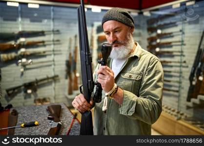 Bearded male hunter buying optical sight for rifle in gun store. Weapon shop interior, ammo and ammunition assortment, firearms choice, shooting hobby and lifestyle. Bearded hunter buying optical sight in gun store