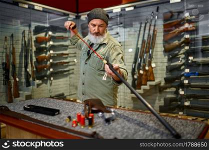 Bearded male hunter cleaning ramrod in gun store. Weapon shop interior, rifles and ammunition assortment, firearms choice, shooting hobby. Bearded male hunter choosing ramrod in gun store