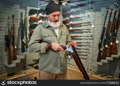 Bearded male hunter loads hunting rifle in gun store. Weapon shop interior, ammo and ammunition assortment, firearms choice, shooting hobby and lifestyle. Bearded male hunter loads rifle in gun store
