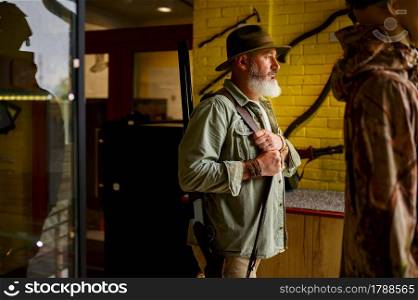 Bearded male hunter tries on hat in gun store. Weapon shop interior, rifles and ammunition assortment, firearms choice, shooting hobby and lifestyle. Bearded male hunter tries on hat in gun store