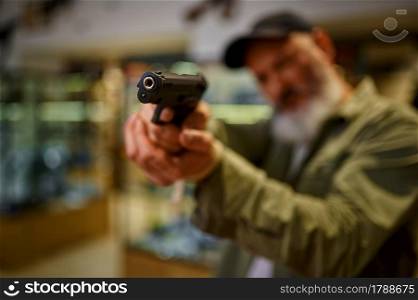 Bearded man aims a pistol in gun store. Weapon shop interior, ammo and ammunition assortment, firearms choice, shooting hobby and lifestyle, self protection. Bearded man aims a pistol in gun store