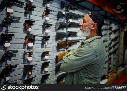Bearded man choosing pistol at the showcase in gun store. Weapon shop interior, ammo and ammunition assortment, firearms choice, shooting hobby and lifestyle, self protection. Man choosing pistol at the showcase in gun store