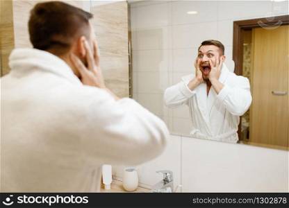 Bearded man frightened by his appearance at the mirror in bathroom, morning hygiene. Male person at the sink performs skin and body treatment procedures. Bearded man frightened by his appearance, hygiene