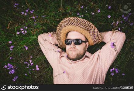 Bearded man in a straw hat and sunglasses rest on the grass among the blue bellflowers. Closeup, space for copy, vintage film filter.. Man Rest On The Grass And Bellflowers