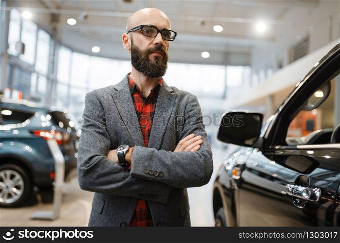 Bearded man in glasses poses at the pickup truck in car dealership. Customer in vehicle showroom, male person buying transport, auto dealer business. Bearded man poses at pickup truck, car dealership