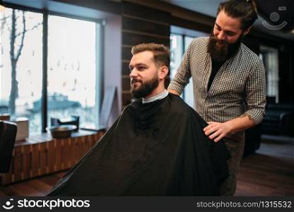 Bearded man in salon cape at the barbershop, barber prepares to cut