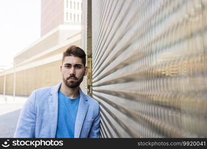Bearded man in urban background wearing casual clothes while leaning on a wall and looking at camera