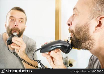 Bearded man looking at himself in mirror trimmng, shaving his beard using electric timmer razor.. Man shaving trimming his beard