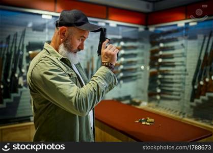 Bearded man poses with black pistol in gun store. Weapon shop interior, ammo and ammunition assortment, firearms choice, shooting hobby and lifestyle, self protection. Bearded man poses with black pistol in gun store