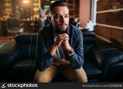 Bearded man sitting on a couch at the barbershop. Hairdressing salon interior on background. Bearded man sitting on a couch at the barbershop