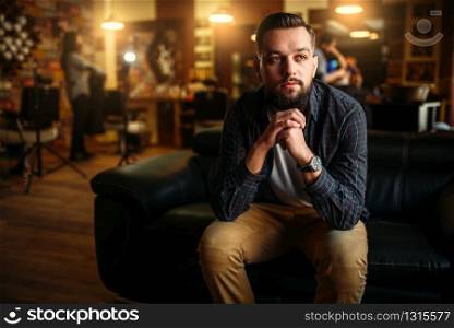 Bearded man sitting on a couch at the barbershop. Hairdressing salon interior on background