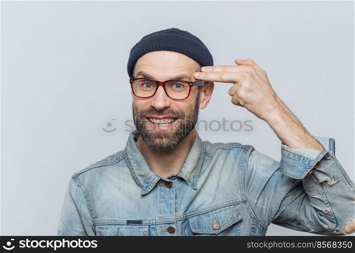 Bearded middle aged man with blue eyes has fun, foolishes and makes suicide gesture, has happy look, wears stylish modern clothing, isolated over grey background. People, positive expressions