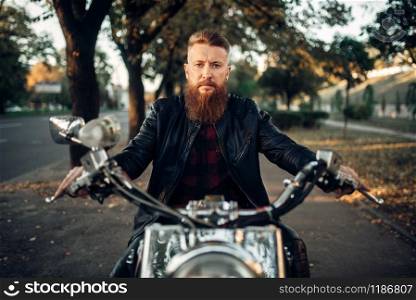 Bearded motorcyclist in leather jacket sitting on classical chopper, front view. Vintage bike, rider on motorcycle