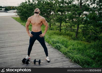 Bearded muscular man keeps hands on waist, raises head and breathes fresh air, takes break after doing workout, trains with barbells. Professional weightlifter works out in nature. Exercising concept