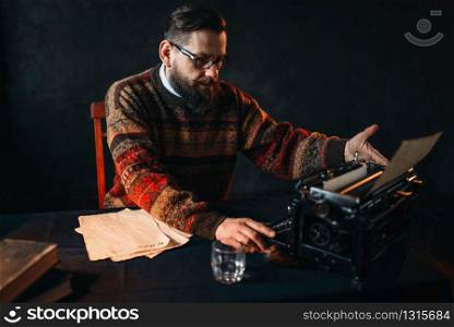 Bearded poet in glasses typing on retro typewriter. Books on the desk with vintage dark blue table cloth. Bearded poet in glasses typing on typewriter