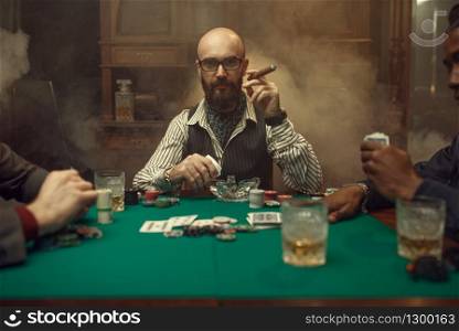 Bearded poker player with cigar, casino. Games of chance addiction. Man leisures in gambling house. Cards, chips and whiskey on gaming table with green cloth. Bearded poker player with cigar, casino
