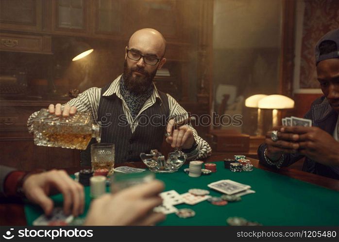 Bearded poker player with cigar pours whiskey, casino. Games of chance addiction. Man leisures in gambling house. Cards and chips on gaming table with green cloth