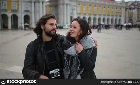 Bearded smiling man hugging girlfriend and pointing at distance. Cheerful tourists with photo camera walking on city square. Travel concept