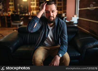 Bearded stylish man waiting on black leather couch at the hairdressing salon. Barber shop interior on background