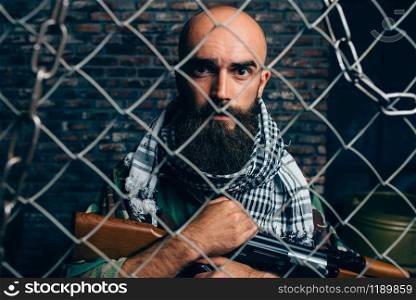 Bearded terrorist in uniform against metal grid, male mojahed. Terrorism and terror, soldier in khaki camouflage, brick wall on background. Bearded terrorist in uniform against metal grid