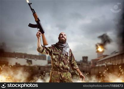 Bearded terrorist with rifle in hands stands in explosion and fire. Terrorism and terror, soldier in khaki camo. Terrorist with rifle stands in explosion and fire