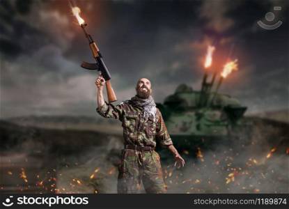Bearded terrorist with rifle in hands stands in explosion and fire. Terrorism and terror, soldier in khaki camouflage. Terrorist with rifle stands in explosion and fire