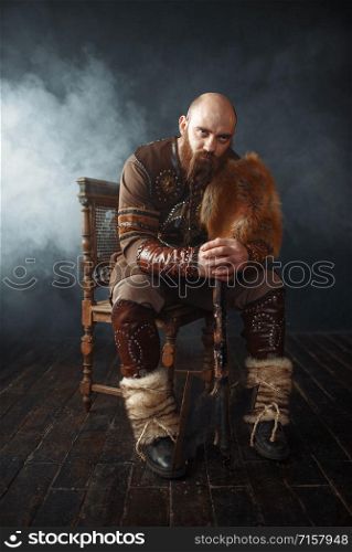 Bearded viking with axe dressed in traditional nordic clothes sitting on chair, barbarian image. Ancient warrior in smoke on dark background