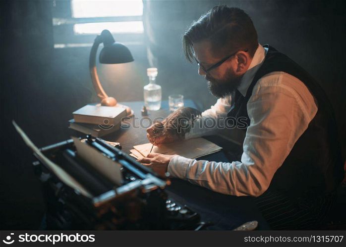 Bearded writer in glasses writes novel with a feather in a smoky room. Retro typewriter, crystal decanter, books and vintage lamp on the desk. Writer in glasses writes novel with a feather