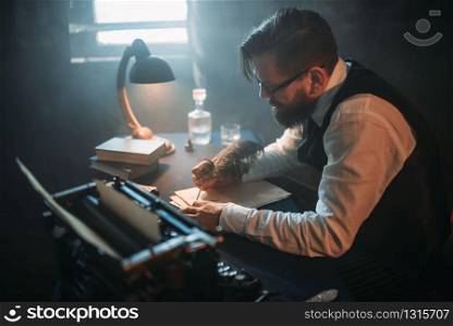 Bearded writer in glasses writes novel with a feather in a smoky room. Retro typewriter, crystal decanter, books and vintage lamp on the desk. Writer in glasses writes novel with a feather