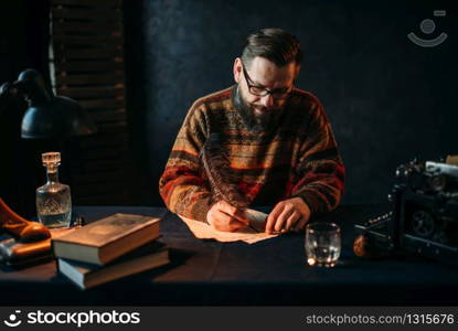 Bearded writer in glasses writes with a feather. Retro typewriter, crystal decanter, books and vintage lamp on the desk. Bearded writer in glasses writes with a feather