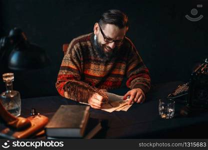 Bearded writer in glasses writes with a feather. Retro typewriter, crystal decanter, books and vintage lamp on the desk. Bearded writer in glasses writes with a feather