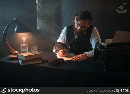 Bearded writer in glasses writes with a feather. Retro typewriter, crystal decanter, books and vintage lamp on the desk. Bearded writer in glasses writes with a feather
