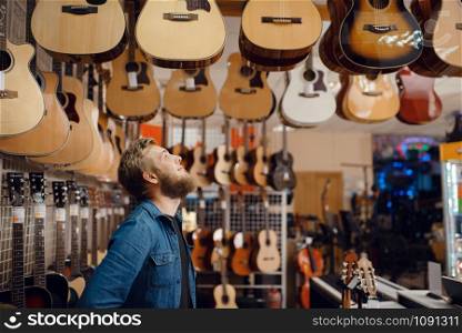 Bearded young guy choosing acoustic guitar in music store. Assortment in musical instruments shop, male musician buying equipment. Young guy choosing acoustic guitar in music store