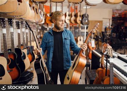 Bearded young guy choosing acoustic guitar in music store. Assortment in musical instruments shop, male musician buying equipment. Young guy choosing acoustic guitar in music store