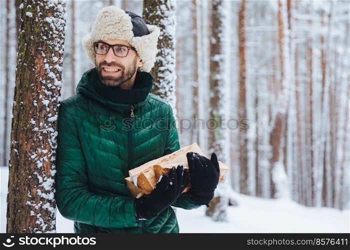 Bearded young male wears warm hat and green jacket, holds pile of firewood, looks happily aside, notices something, likes winter and frosty weather. Dreamful attractive man spends time in forest