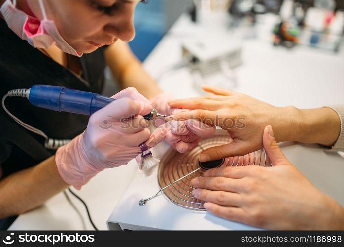 Beautician in gloves with polishing machine treats nails of female client, manicure in beauty salon. Manicurist doing hands care cosmetic procedure