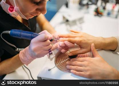 Beautician in gloves with polishing machine treats nails of female client, manicure in beauty salon. Manicurist doing hands care cosmetic procedure