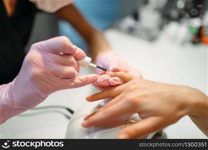 Beautician in pink gloves applying nail varnish to female client, manicure in beauty salon. Manicurist doing hands care cosmetic procedure. Beautician applying nail varnish to female client